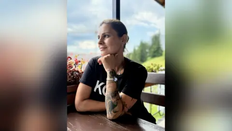Thoughtful woman in a black t-shirt sitting at a wooden table by a window with a scenic view.