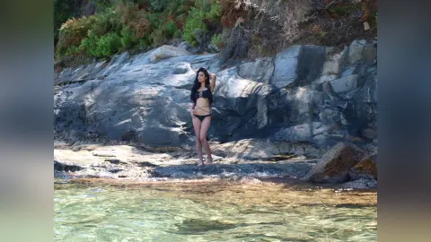 Woman in black bikini standing on rocky coastline by clear turquoise ocean water under bright sunlight.