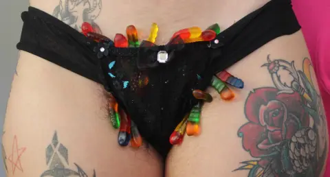 Tattooed woman wearing black lace underwear decorated with colorful gummy candies in a playful mood.