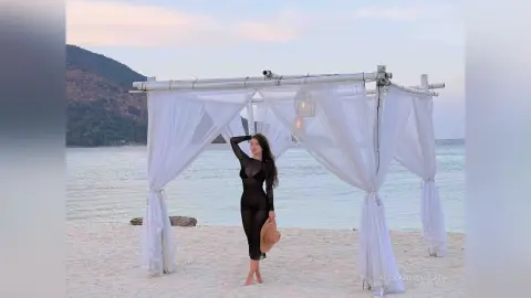 Elegant woman in a black sheer maxi dress standing under a white canopy on a serene tropical beach at sunset.