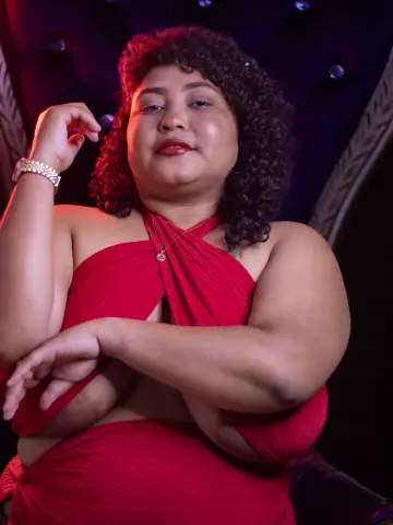 Curvy woman in a glamorous red halter dress posing confidently in a moody, dimly lit setting.
