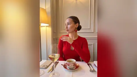 JessicaMelori wears a long-sleeved red top while sitting at a table with a glass of white wine and a cup of coffee in a bright room.