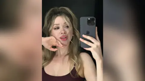 CassieArmas poses for a mirror selfie, wearing a maroon tank top and showing her long blonde hair and face.