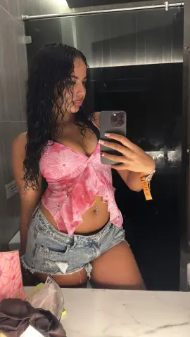 JasmineDesire22 poses in a pink patterned wrap top and denim cut-off shorts, showing her long dark wavy hair and navel piercing in a mirror selfie.