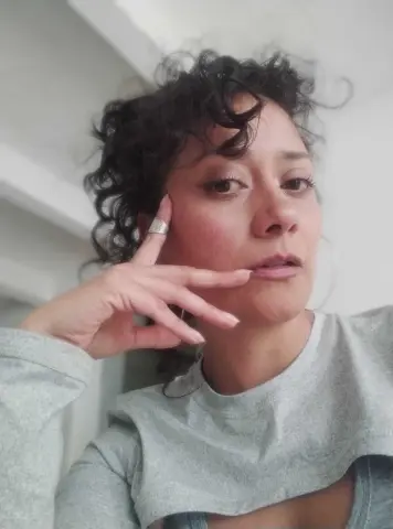 Close-up portrait of a woman with curly hair wearing a grey long-sleeve shirt in a bright, minimalist setting.