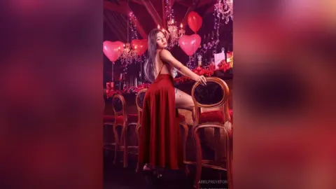 Elegant woman in a red satin dress posing with heart balloons in a romantic, dimly lit lounge setting.