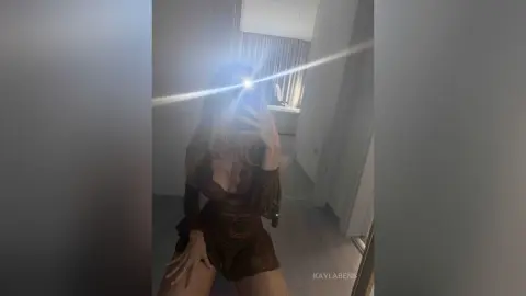 Young woman in a dark brown patterned romper posing for a mirror selfie in a modern bedroom.
