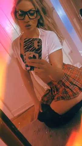 Young woman in white t-shirt and red plaid skirt taking a mirror selfie in a warm indoor setting.