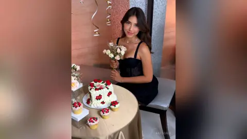 Smiling woman in a black dress sitting by a white floral cake at a celebratory indoor party.