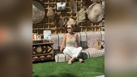 Stylish woman in white pants and beige tank top sitting on a patterned outdoor sofa in a boho garden setting.