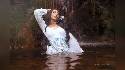 Ethereal woman in a white flowing dress and blue corset relaxing in a forest waterfall pool.
