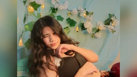 Young woman in a black top winking playfully against a dreamy floral and fairy light background.