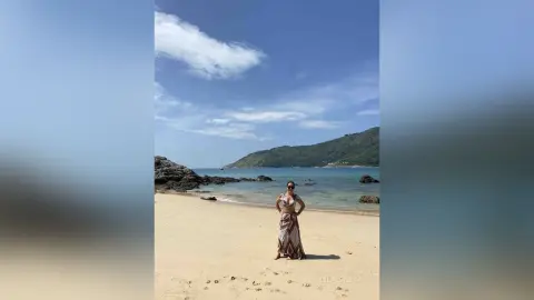 Stylish woman in a patterned maxi dress standing on a sunny tropical beach with ocean waves and mountains.