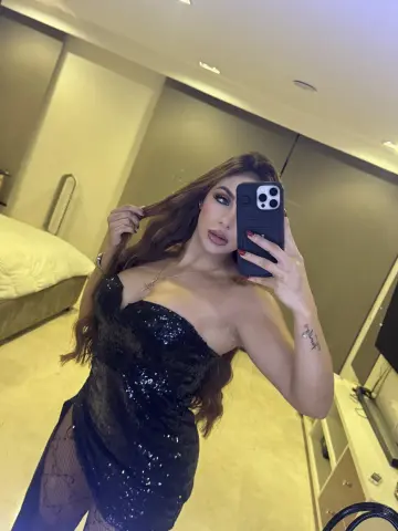 Woman in a black sequin cocktail dress taking a glamorous mirror selfie in a modern bedroom.
