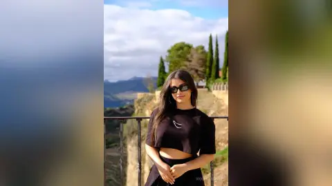 Young woman in black crop top and sunglasses posing outdoors against a scenic mountain landscape.