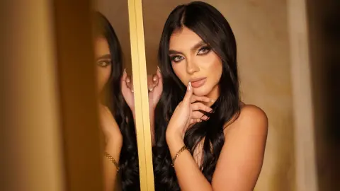 Glamorous woman with long dark hair and smoky eye makeup posing in a warm, golden indoor setting.