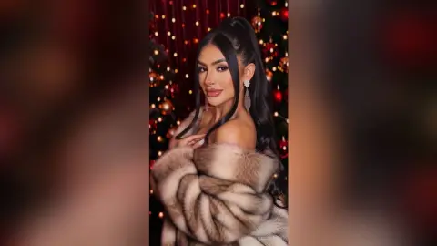 Glamorous woman in a luxury faux fur coat posing against a festive Christmas tree background with warm lights.