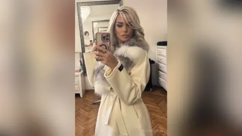 Blonde woman in a stylish cream coat with a grey faux fur collar taking a selfie in a bright room.