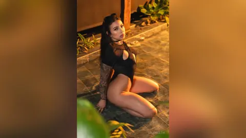 MilaWillow sits on a tiled patio in a black bodysuit, showing off her long dark hair and tattoos.