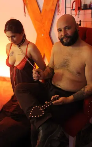 A woman in a red slip dress holding a chain next to a shirtless man in dark pants in a warm, moody setting.
