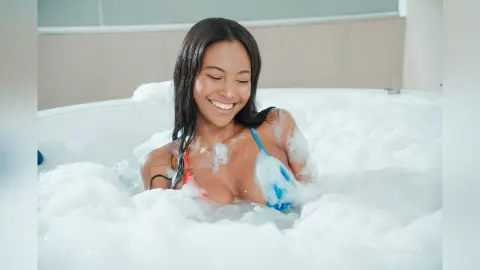 Smiling young woman in a blue bikini relaxing in a bubbly white foam hot tub for a peaceful spa experience.