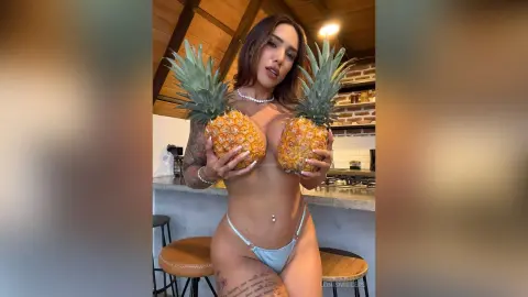 Brunette woman in light blue bikini holding pineapples in a modern kitchen setting.