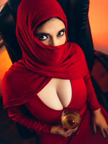 Woman in a red hijab and matching dress sitting in a chair holding a glass of sparkling drink.