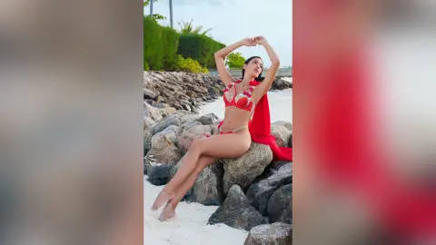 Elegant woman in a red bikini sitting on beach rocks under a sunny sky.