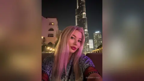 Blonde woman in a blue patterned top posing at night with the Burj Khalifa skyline in the background.