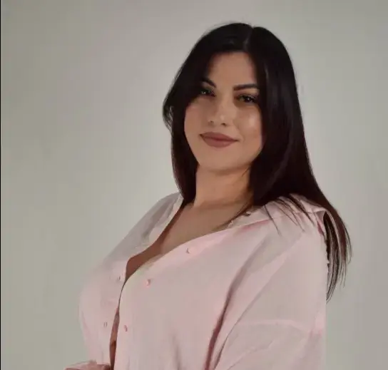 Smiling woman with long dark hair wearing a pink blouse against a neutral studio background.