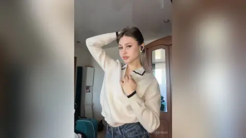 Young woman in a cream long-sleeve shirt and blue jeans posing in a bright modern room.