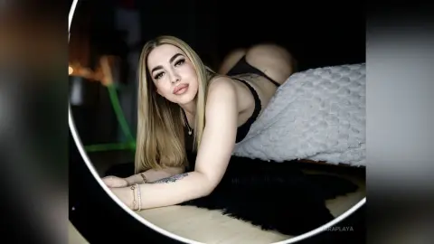 ZaraPlaya poses on her stomach, showing her blonde hair and bare back while wearing black lingerie in a dimly lit room.