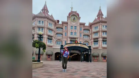 Young woman in a blue patterned shirt and olive pants standing before the Disneyland Park entrance building.