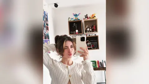 Young woman in a white cable knit cardigan taking a playful selfie in a room filled with anime figurines.