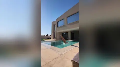 EvelynMills stands in a clear blue swimming pool next to a modern tan house under a bright blue sky.