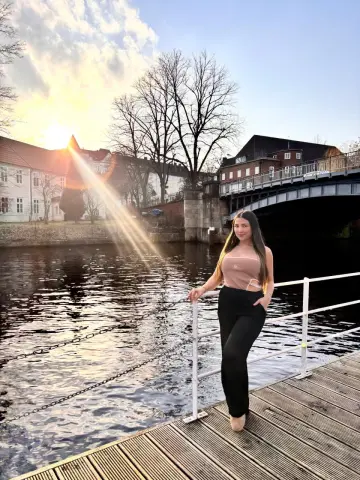 Woman in a beige top and black trousers standing on a wooden pier by a river at sunset.
