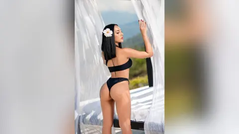 Elegant woman in black bikini with white flower hair accessory posing by sheer curtains in a sunny outdoor setting.