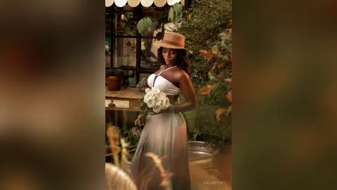 Elegant woman in a white summer dress and straw hat holding white roses in a lush garden setting.