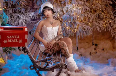 Tattooed woman in white corset sitting on a park bench in a snowy winter wonderland with a red Santa Mail box.