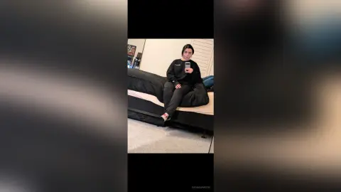 Young woman in a black sweatshirt sitting on a bed in a cozy bedroom setting.