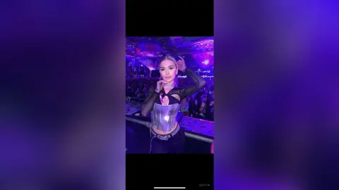DaisyLin poses in a black mesh top and silver corset, running her hands through her blonde hair under purple club lighting.