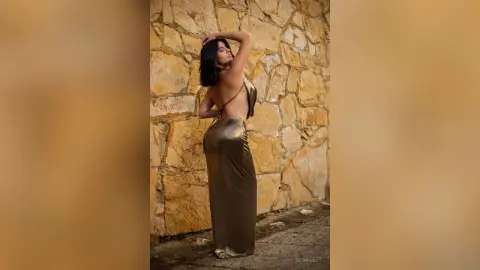 Elegant woman in a shimmering gold satin backless gown posing against a rustic stone wall in warm sunlight.