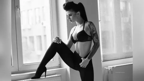 Elegant woman in black lingerie and tights posing by a window in a moody black and white indoor setting.