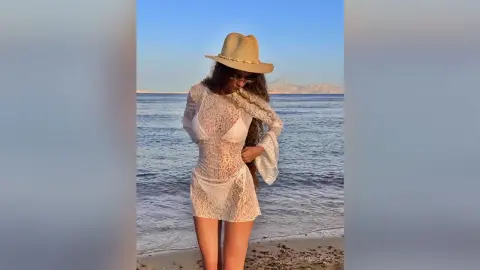 Woman in white lace mini dress and straw hat posing on a sunny beach at sunset.