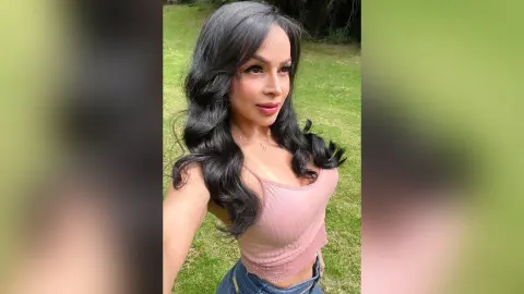 Smiling woman with long wavy black hair wearing a pink tank top in a sunny green garden setting.