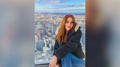 Smiling woman in a black puffer jacket and jeans overlooking a sunny city skyline from a high-rise balcony.