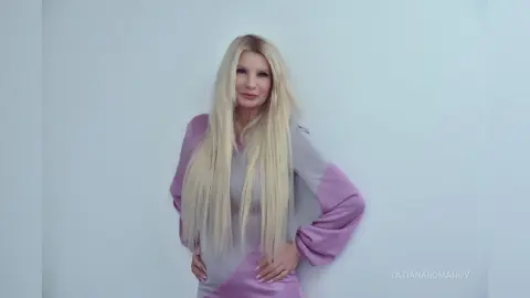Blonde woman in a stylish lavender long-sleeve dress posing against a minimalist white background with a calm mood.