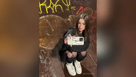 Young woman in a black sweatshirt taking a selfie against a dark graffiti wall in a moody urban setting.