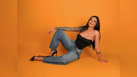 Smiling woman in black tube top and blue jeans sitting against an orange background.
