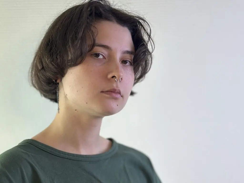 Young woman with short dark hair and nose ring wearing a green t-shirt against a white wall background.